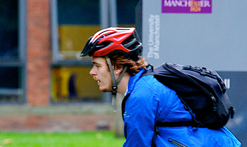 A male student on a bicycle with the University logo behind him