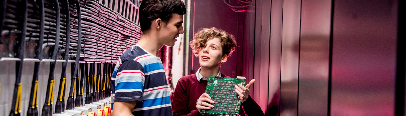 Two students in discussion about a computer circuit board in a corridor