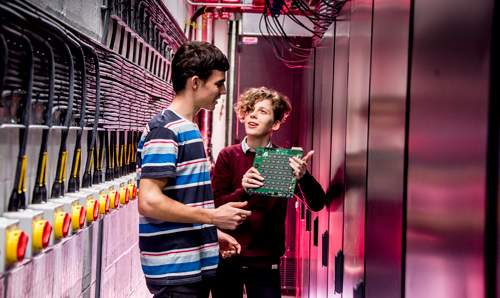 Two students in discussion about a computer circuit board in a corridor